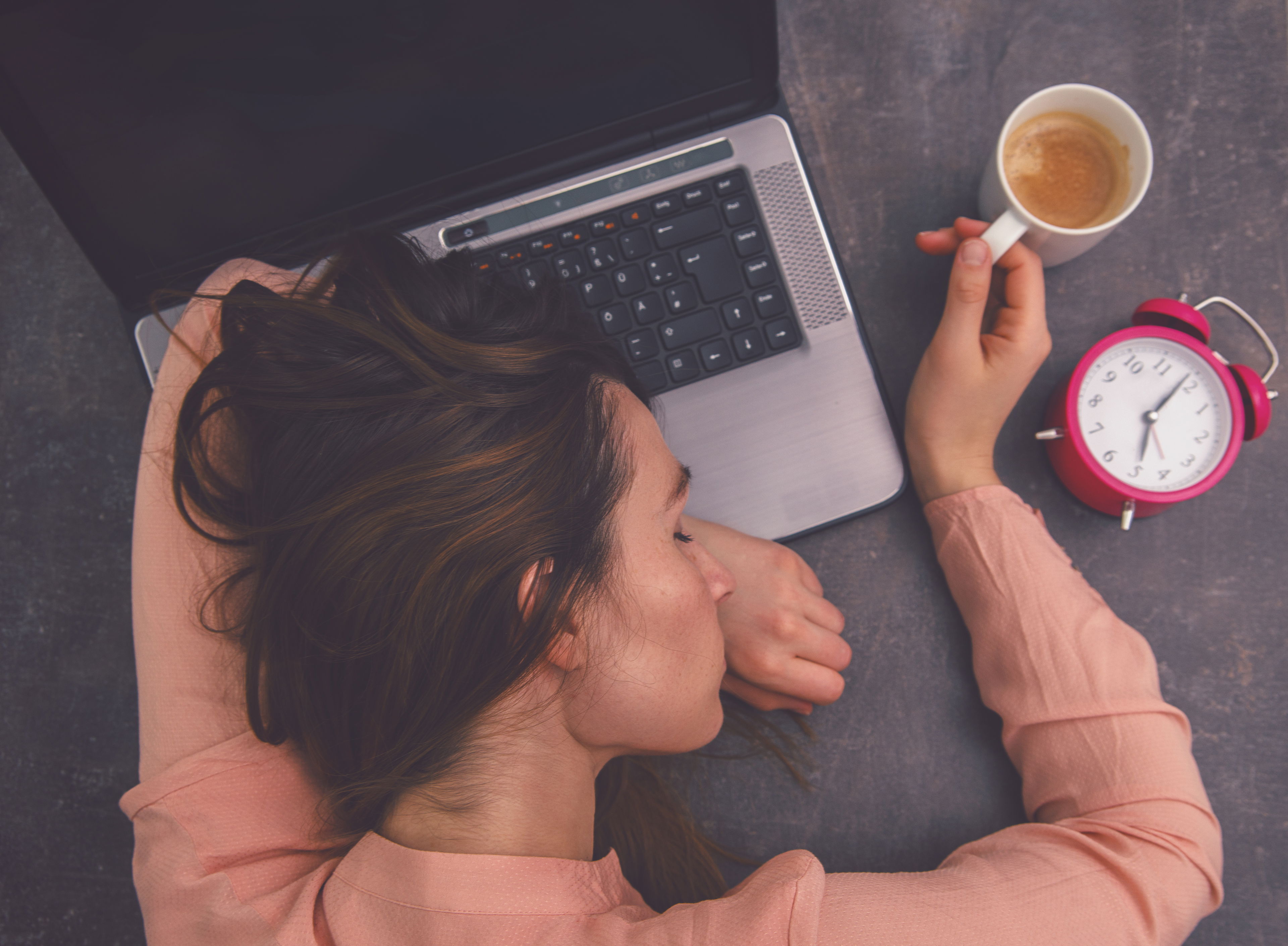Die Macht des Powernaps – Erholung im Alltag Frau schläft erschöpft mit dem Kopf auf dem Laptop, in der Hand eine halbvolle Kaffeetasse, daneben ein Wecker – Symbolbild für Powernapping und Müdigkeit am Arbeitsplatz.