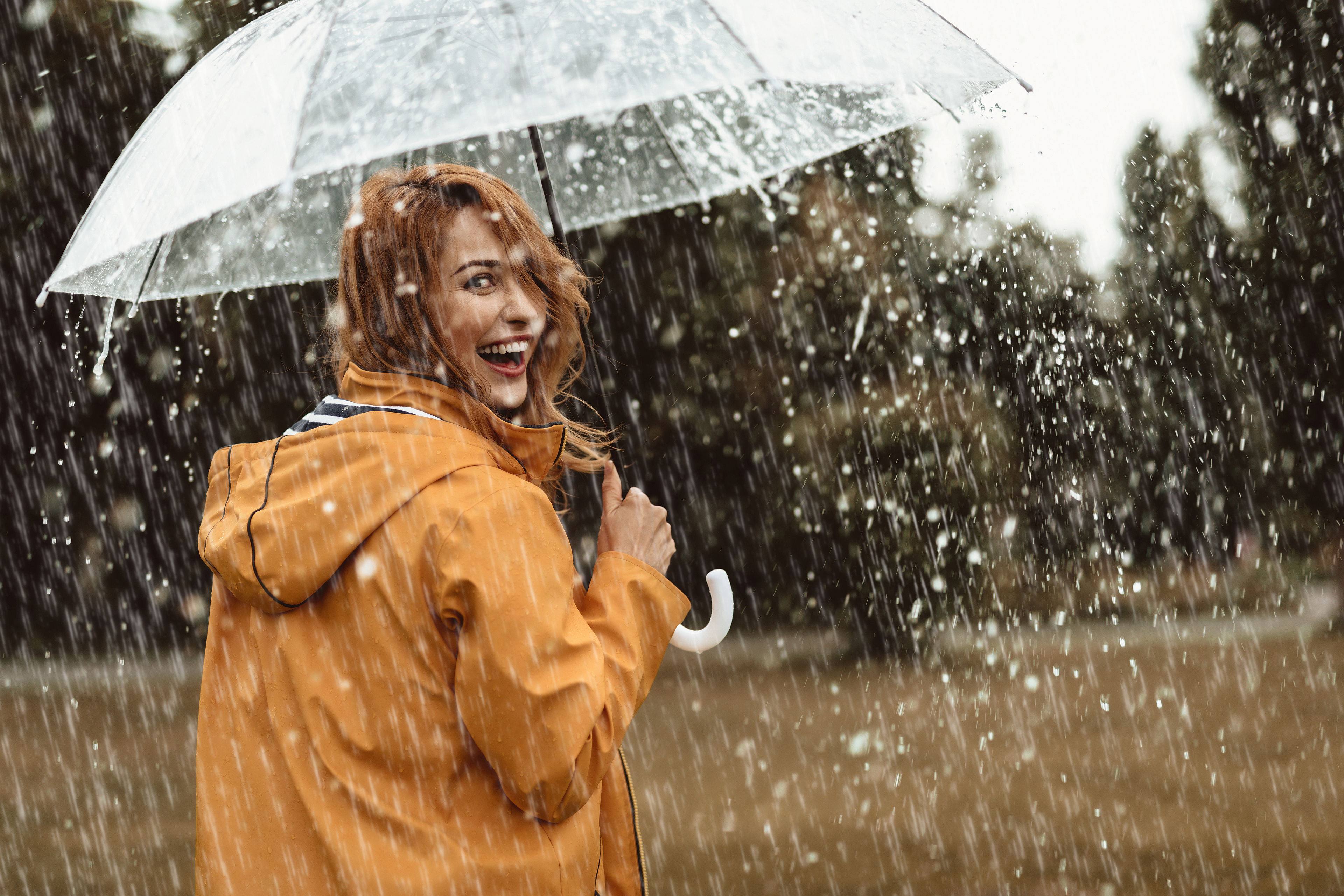 Lachende Frau in gelber Regenjacke unter einem durchsichtigen Regenschirm im Regen – Symbol für Widerstandskraft und Wohlbefinden durch präventive CBD-Anwendung.