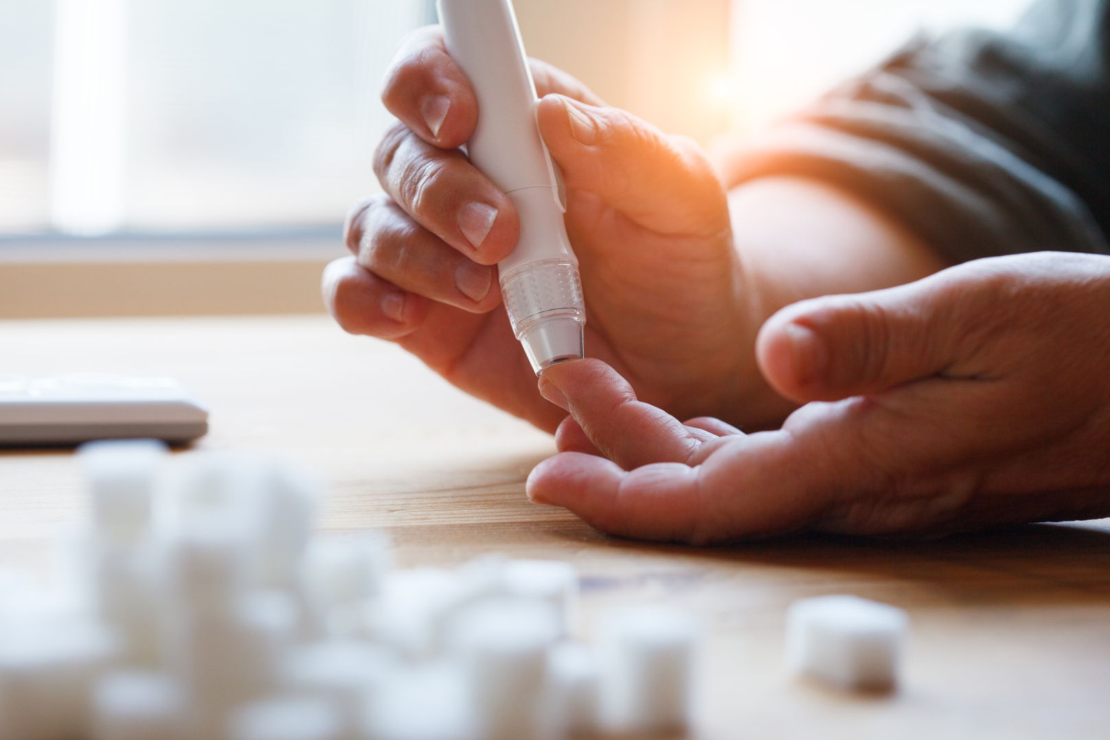 CBD bei Diabetes – Unterstützung für ein stabiles Leben Nahaufnahme einer Hand, die mit einem Blutzuckermessgerät den Blutzucker misst, auf einem Tisch mit Würfelzucker im Vordergrund.