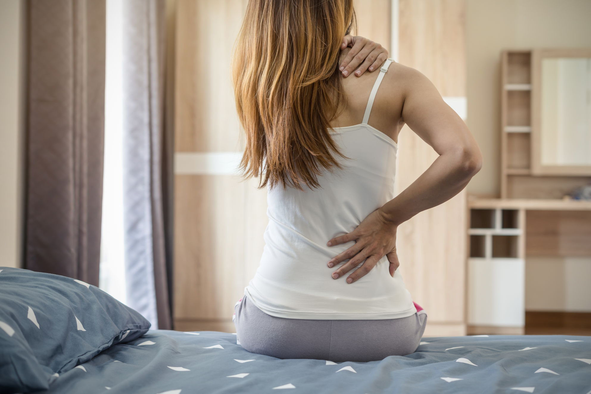 CBD bei Fibromyalgie – Wenn der ganze Körper schmerzt Eine Frau sitzt auf einem Bett und hält sich mit schmerzverzerrter Haltung sowohl den unteren Rücken als auch die Schulter – Symbolbild für chronische Ganzkörperschmerzen.