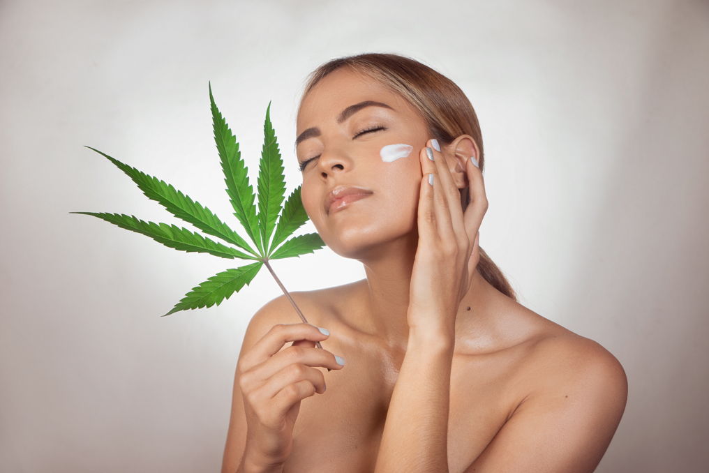CBD in der Hautpflege – natürlicher Anti-Aging-Booster mit Cannabidiol Junge Frau trägt Creme auf ihr Gesicht auf und hält ein großes Hanfblatt – Symbolbild für CBD-Kosmetik und Anti-Aging-Pflege
