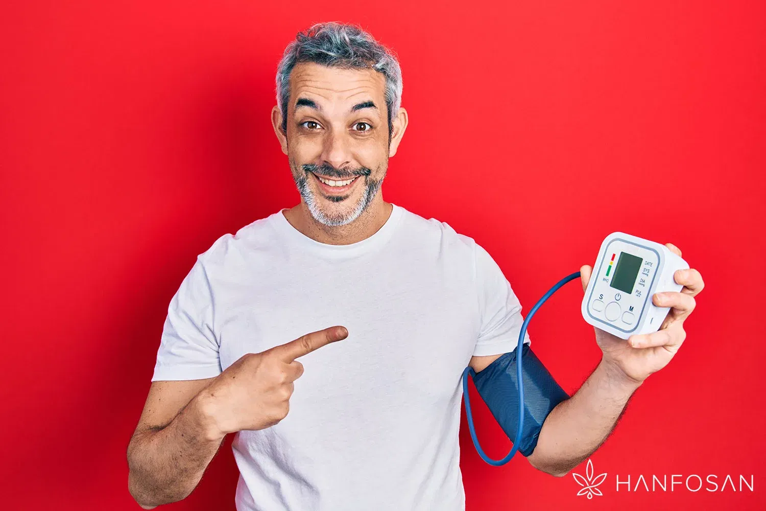 Fröhlicher Mann in weißem T-Shirt misst mit einem digitalen Blutdruckmessgerät seinen Blutdruck, vor rotem Hintergrund, unten rechts mit Hanfosan-Logo