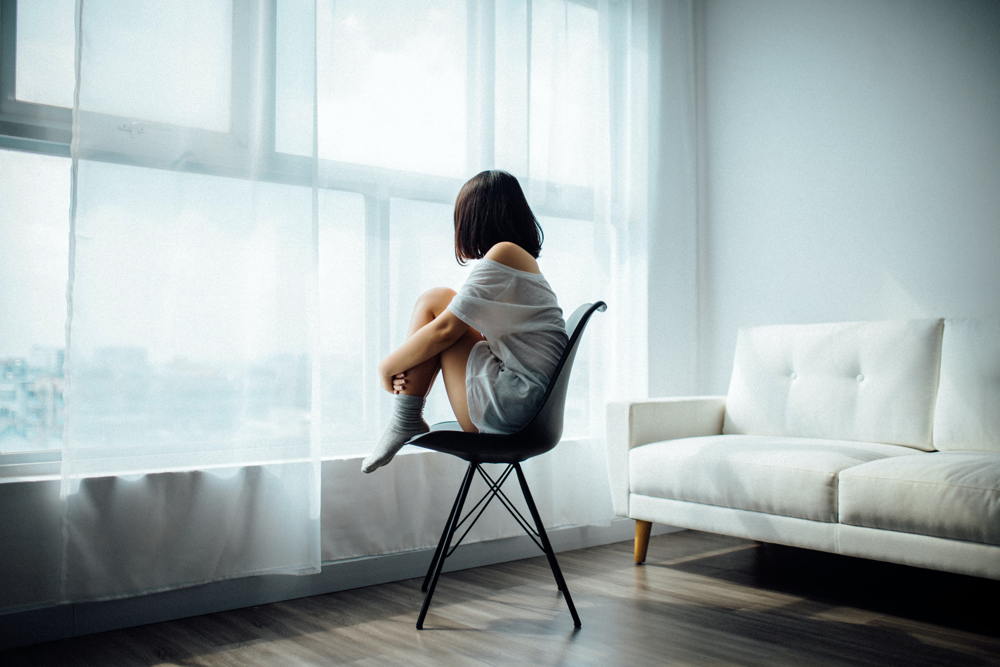 CBD bei Depressionen – Hoffnung in dunklen Zeiten Frau sitzt allein in einem hellen Raum auf einem Stuhl und blickt gedankenversunken aus dem Fenster – Symbolbild für Depression, Einsamkeit und emotionale Belastung.