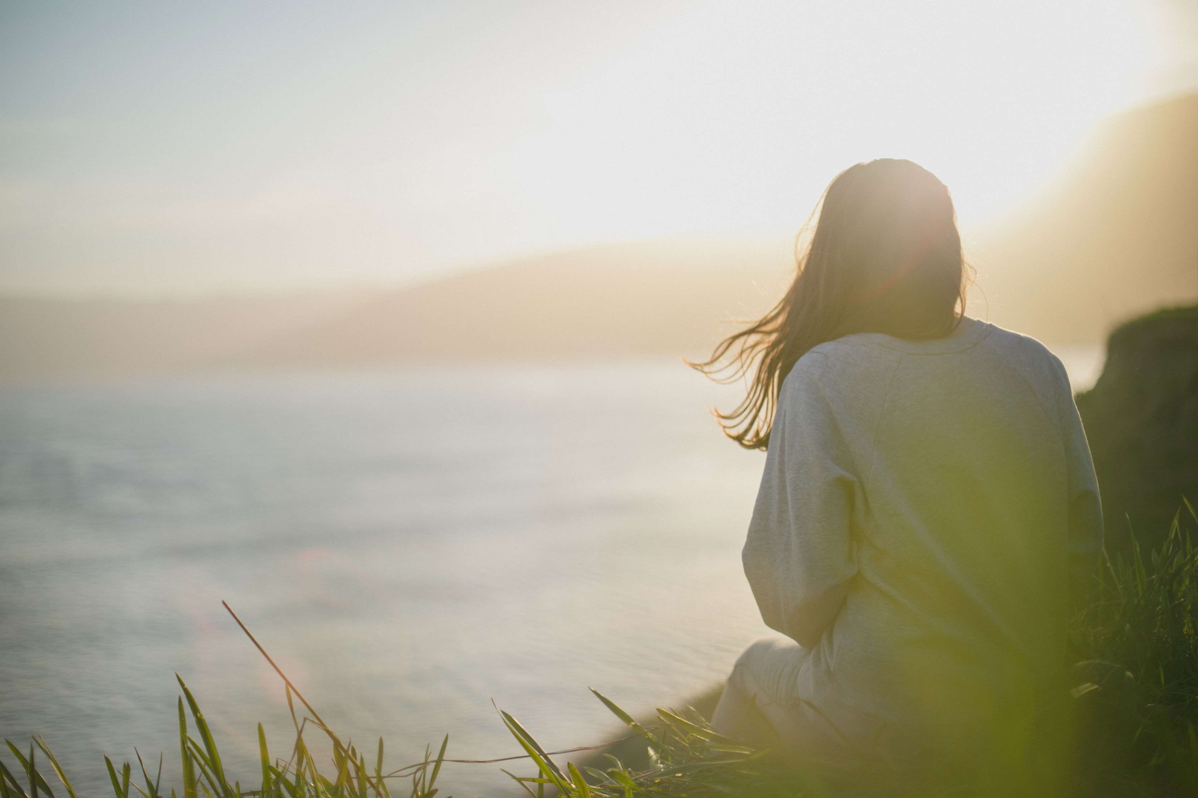 CBD gegen Liebeskummer – Ruhe finden in emotional stürmischen Zeiten Eine Frau sitzt allein im Gras an einer Klippe mit Blick auf ein stilles Gewässer im Sonnenuntergang – Symbolbild für Trauer, Selbstreflexion und emotionale Heilung.