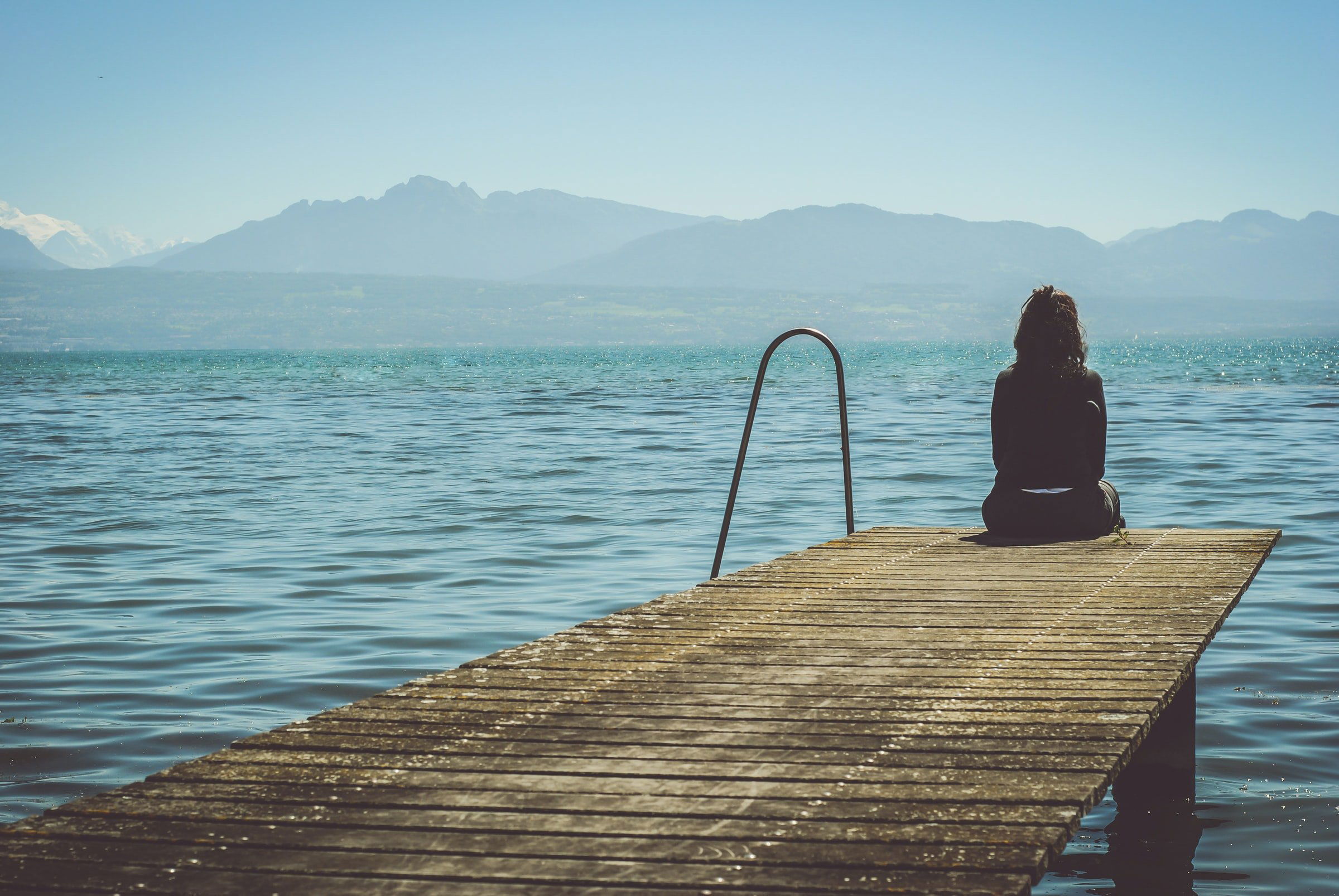 CBD bei psychischen Erkrankungen – Balance finden zwischen den Gedanken Frau sitzt alleine am Ende eines Stegs am See, den Blick auf das Wasser und die Berge gerichtet – symbolisiert Rückzug, Nachdenklichkeit und Hoffnung bei psychischer Belastung.