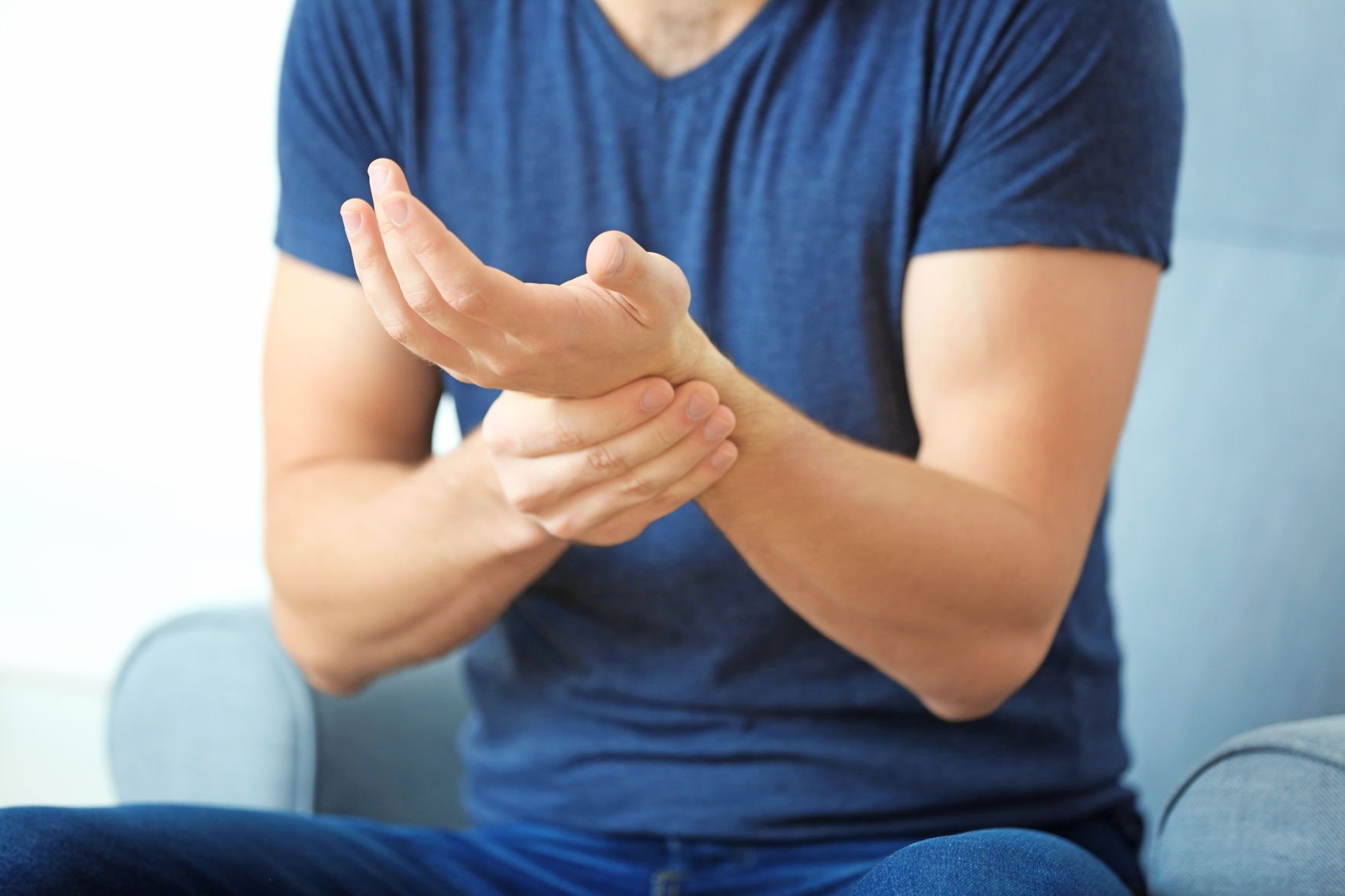 CBD bei Rheuma – Mann hält sich schmerzhaft das Handgelenk Ein Mann in einem blauen T-Shirt sitzt auf einem Sessel und hält sich mit schmerzverzerrtem Gesicht das rechte Handgelenk – typisches Symptom bei rheumatischen Beschwerden.