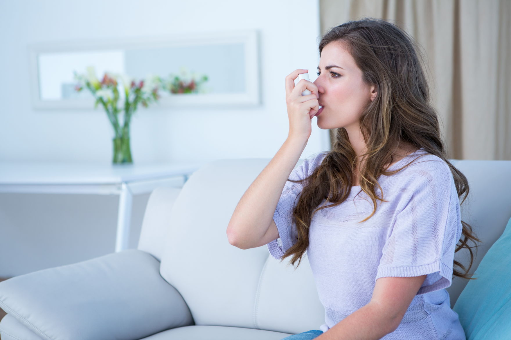 CBD bei Atemwegserkrankungen – natürliche Ergänzung bei Asthma Junge Frau mit Inhalator sitzt auf dem Sofa und benutzt ihr Asthmaspray – Symbolbild für Atembeschwerden und Asthma