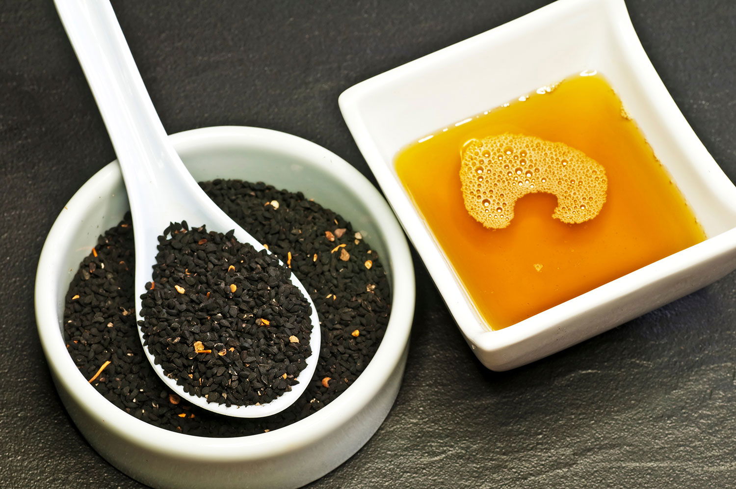 Schwarzkümmelöl und Schwarzkümmelsamen – Naturheilmittel in ihrer reinsten Form Weiße Schale mit Schwarzkümmelsamen und Keramiklöffel neben einer Schale mit goldgelbem Schwarzkümmelöl auf dunklem Untergrund