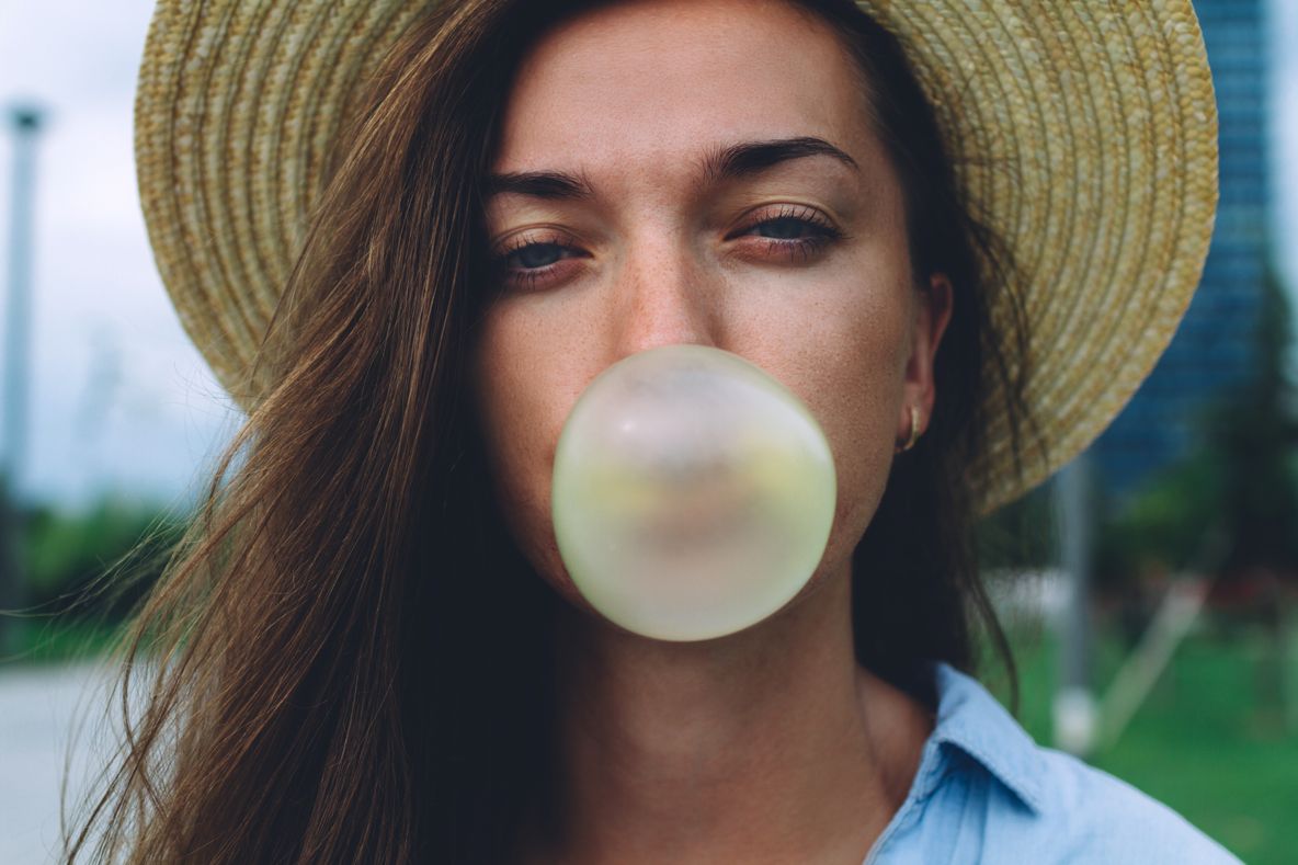 Frau mit Strohhut bläst einen Kaugummiballon – Symbolbild für CBD-Kaugummi als moderne Einnahmeform Frau mit Strohhut bläst einen Kaugummiballon – Symbolbild für CBD-Kaugummi als moderne Einnahmeform