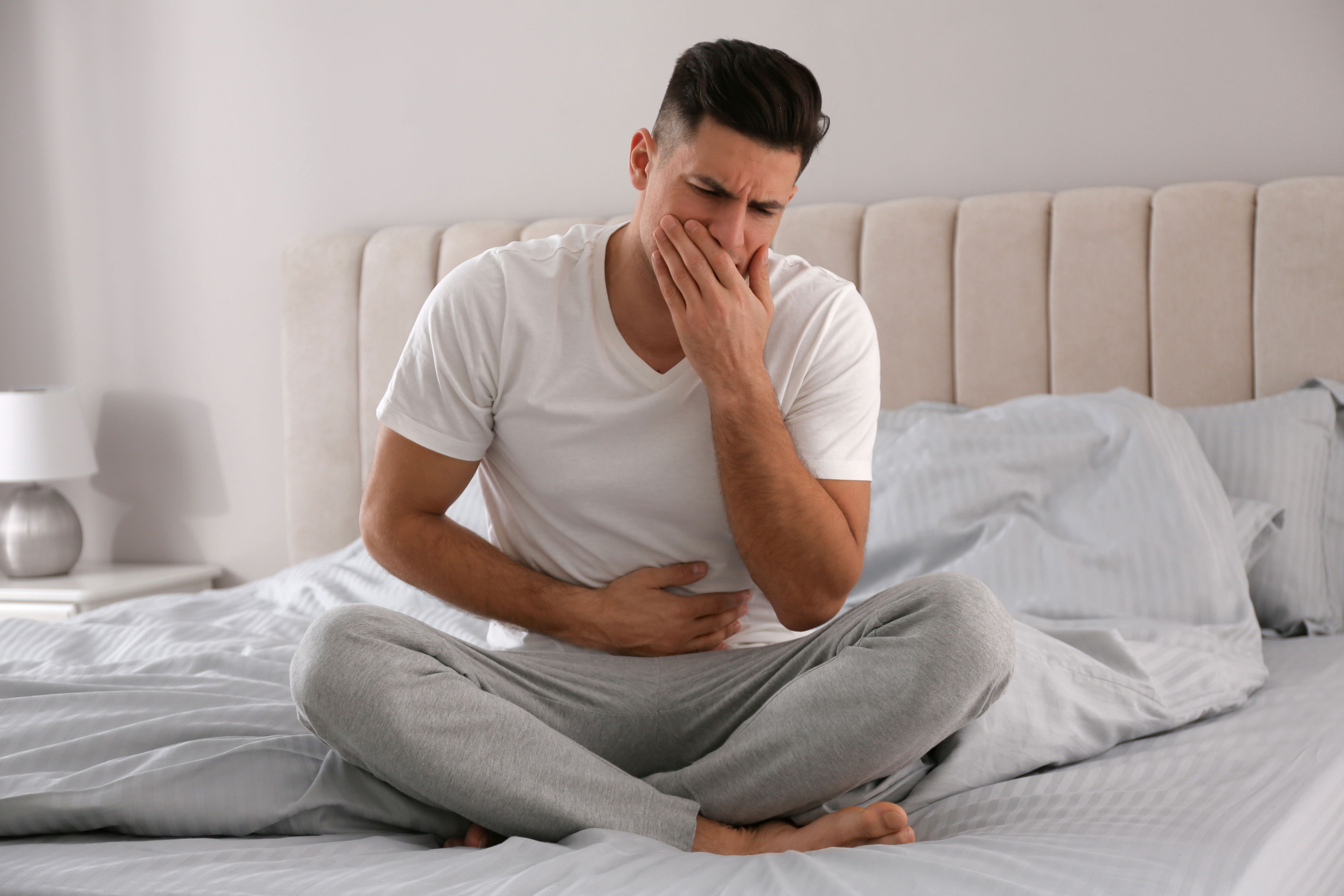 CBD gegen Übelkeit – Linderung auf natürliche Weise Ein junger Mann sitzt im Schneidersitz auf dem Bett, hält sich den Bauch und bedeckt mit der anderen Hand seinen Mund – Ausdruck starker Übelkeit und Unwohlsein.