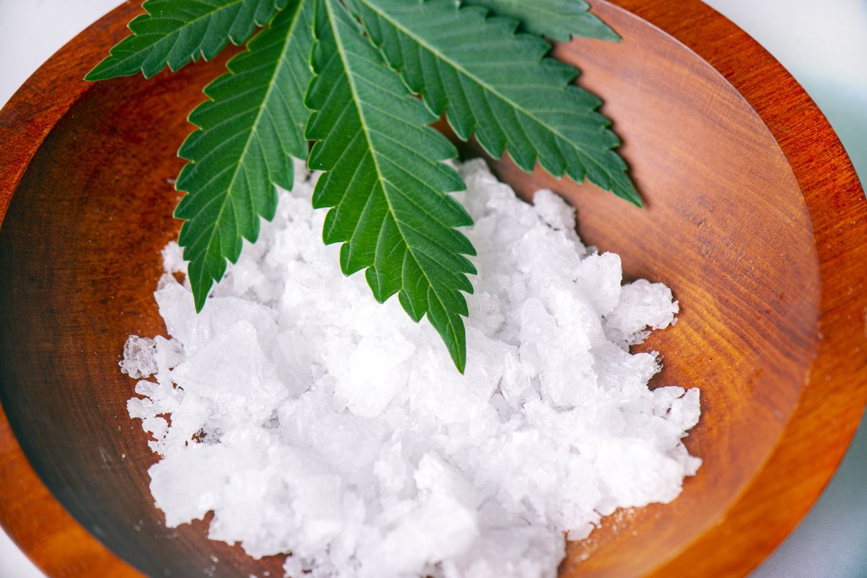 Weiße CBD-Kristalle in einer Holzschale, dekoriert mit einem Hanfblatt – Symbolbild für reines Cannabidiol-Isolat.