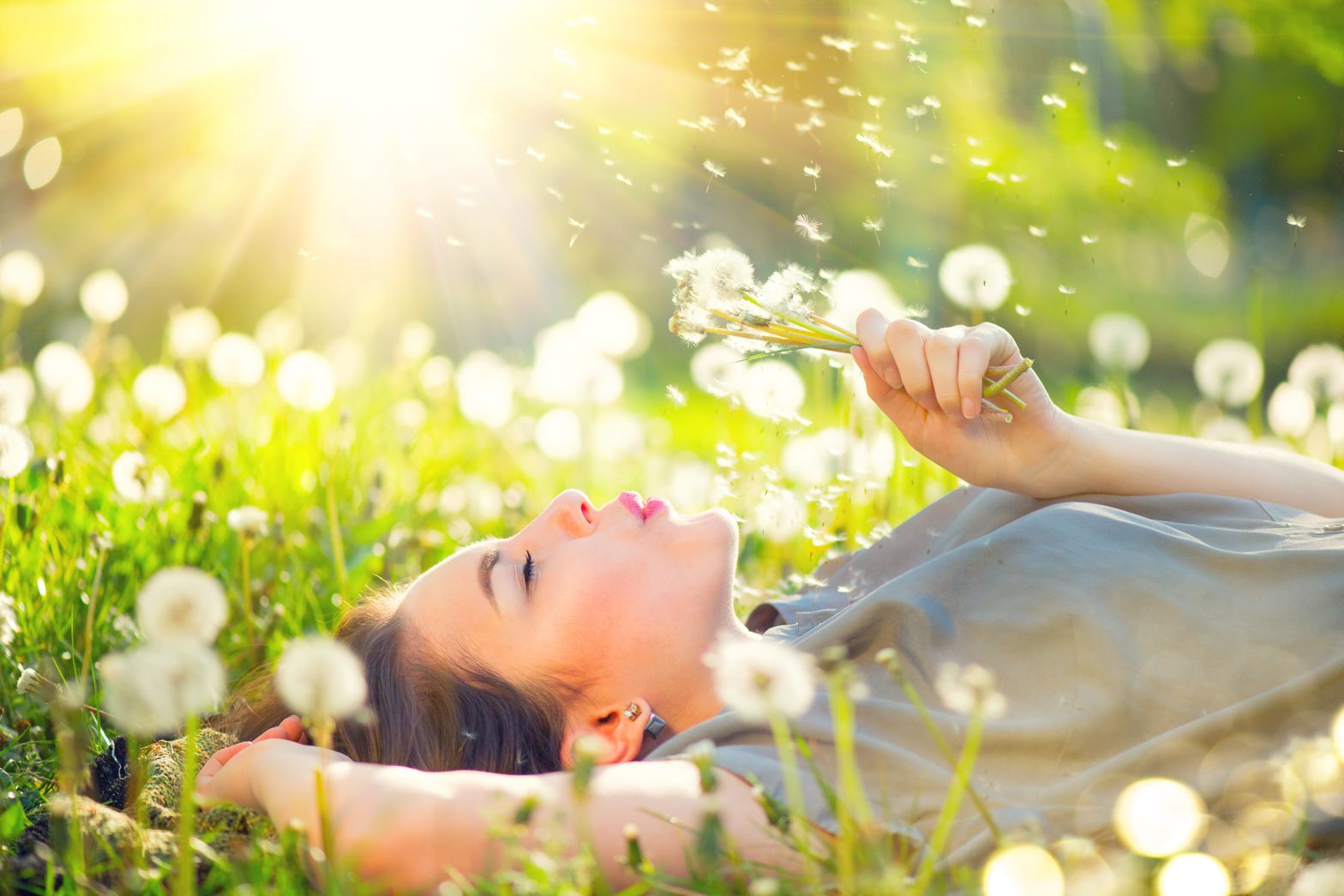CBD bei Heuschnupfen – natürliche Erleichterung in der Allergiezeit Junge Frau liegt entspannt auf einer blühenden Frühlingswiese und pustet in eine Handvoll Pusteblumen – Symbol für Natur, Leichtigkeit und Heuschnupfenzeit.