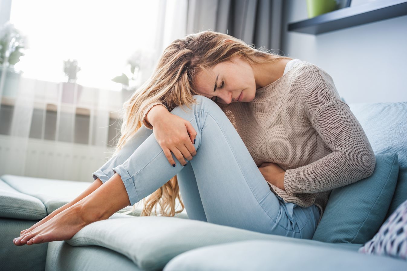 CBD bei Regelschmerzen – Linderung auf natürliche Weise Junge Frau sitzt mit schmerzverzerrtem Gesicht auf dem Sofa, hält sich den Unterbauch – Symbolbild für Menstruationsbeschwerden.